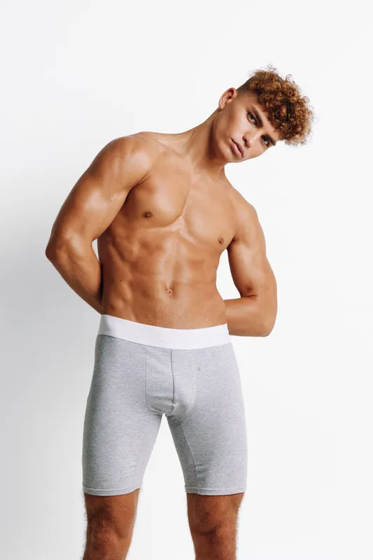 Muscular young man shirtless posing with hands behind back wearing gray boxer briefs against white background in studio. Fitness, bodybuilder and strong male model with defined abs.