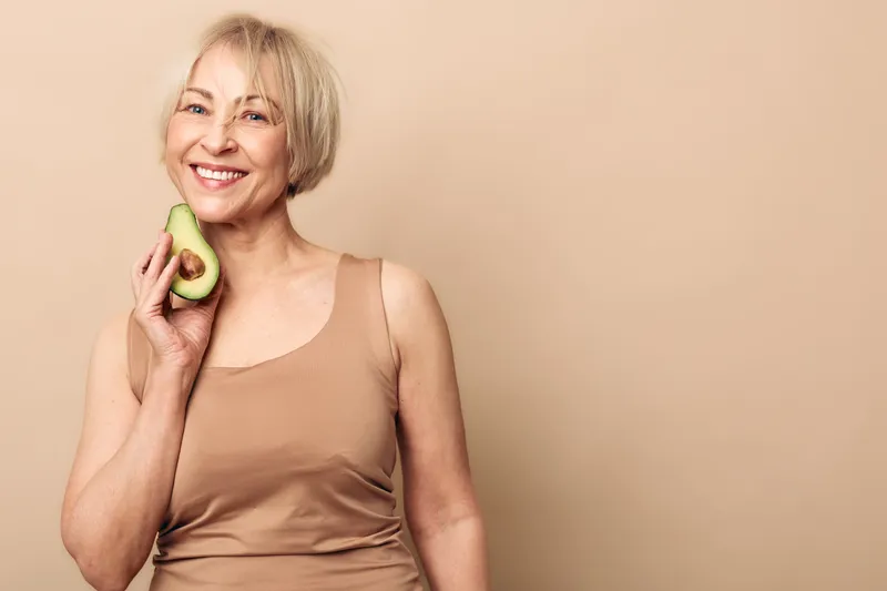 Smiling mature woman holds fresh avocado near face, wearing beige tank top against soft neutral background. Concept of healthy lifestyle, nutrition, natural beauty, and wellness for older adults.