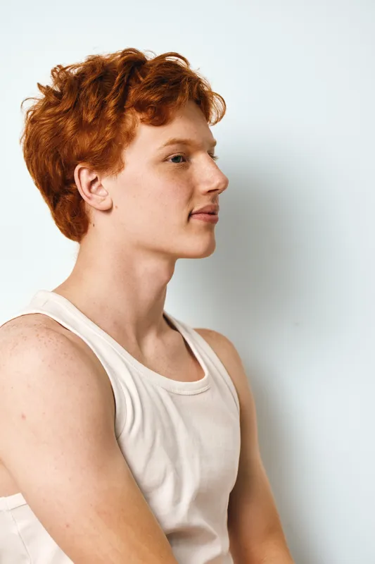 Profile portrait of young redhead man wearing white tank top looking forward with neutral expression on white background. Casual style and natural light with copy space.