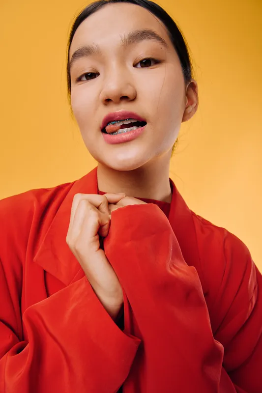 Young woman with braces wearing bright red jacket posing in front of orange background. Closeup portrait of confident female with natural makeup and casual style posing for camera.