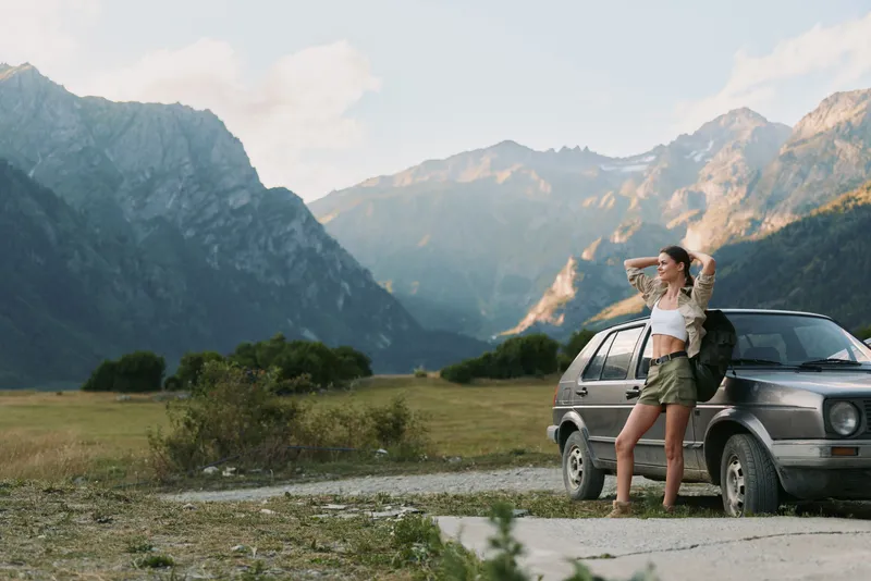 Young woman with backpack stands beside a car, gazing at mountains. Outdoors lifestyle with travel and adventure vibes, illuminated by natural light on a vast scenic landscape.
