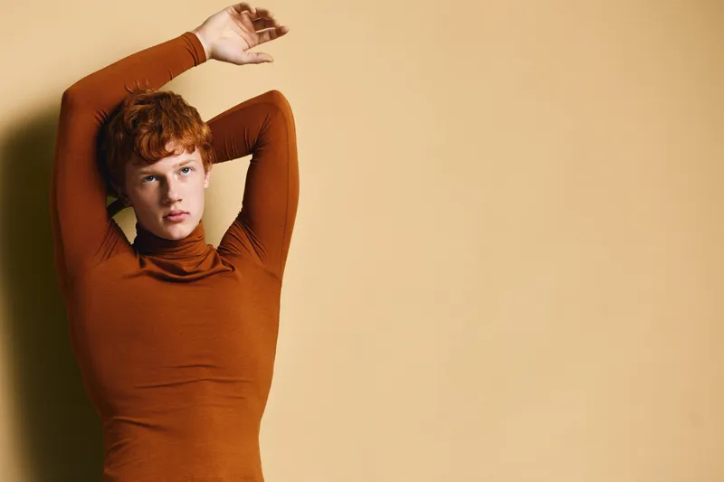 Woman with red hair and pale skin wearing brown long sleeve top posing with hands raised against beige wall in minimal studio setting with soft lighting and shadow.