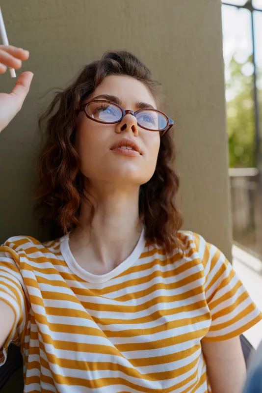 Young woman wearing a yellow striped tee sits near a balcony, thoughtful mood, casual home lifestyle, natural light portrait, relaxed pose, contemporary style.