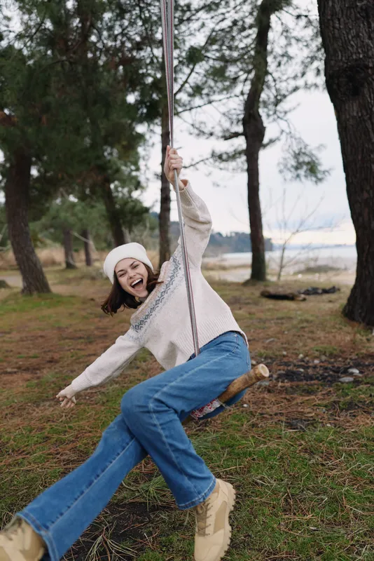 Girl on a swing in a park, outdoor joyful moment with trees and grass, smiling casually in a cozy sweater, jeans and boots, capturing playful motion and carefree childhood energy.