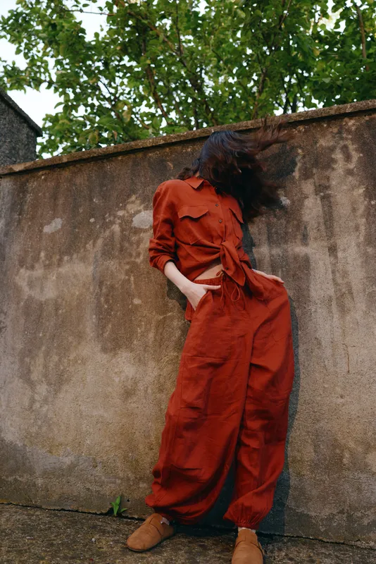 A woman in a rust orange jumpsuit stands leaning against a weathered wall outdoors, hands in pockets, relaxed pose, casual street fashion captured in natural light