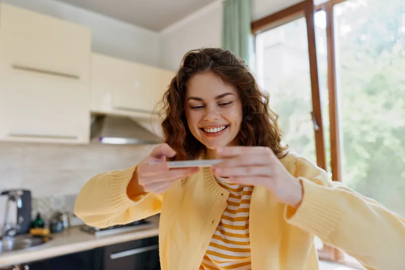 Cheerful woman indoors in a bright kitchen, smiling while checking her phone, casual sweater, warm sunlight streaming through a window, relaxed home lifestyle moment