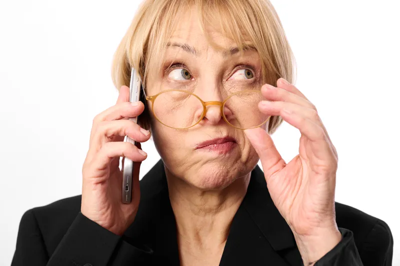 Elderly woman with glasses, phone call, studio portrait, face closeup, adjusting eyewear, calm thoughtful expression, white background, indoor communication