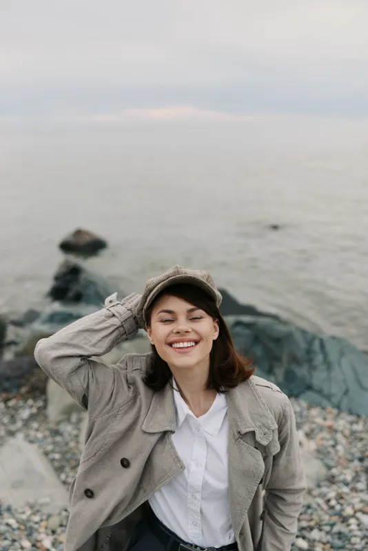 Smiling young woman in casual autumn clothes stands on rocky beach near calm sea. She wears hat and coat, expresses happiness and relaxation in outdoor coastal environment.