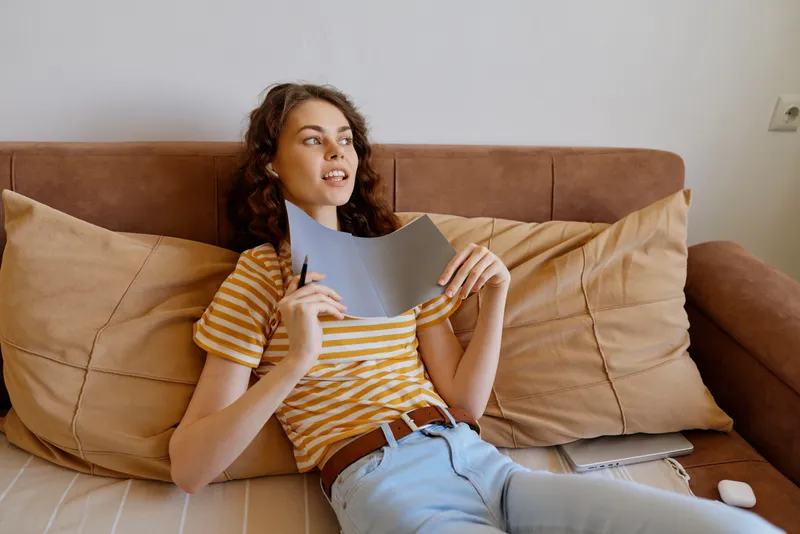 Young woman relaxes at home on a sofa, reading a letter, wearing a striped t shirt and jeans, casual cozy moment, natural light, intimate domestic setting, with soft cushions.
