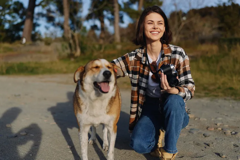 Woman kneels beside a friendly dog on a sunny outdoor park path, capturing memories with a camera and enjoying companionship with her pet on a bright day.