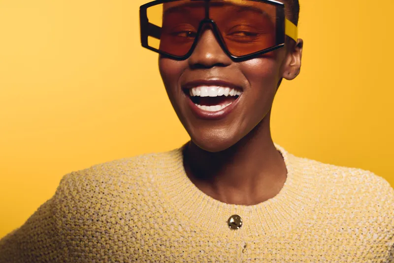 Smiling dark skinned African woman with short hair wearing large brown sunglasses and a beige knitted sweater. Bright yellow background creates a cheerful and fashionable portrait concept.