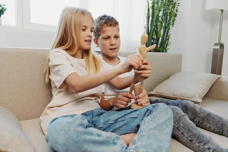 Two children sitting on sofa playing with wooden mannequin toy, enjoying creative activity in bright living room with natural light and casual clothes.