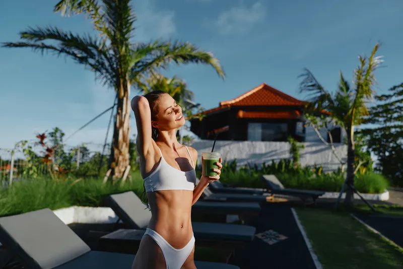 Young woman in a white bikini relaxes by a pool at a tropical resort, sipping a green smoothie and basking in warm sunlight, her relaxed smile conveying wellness and summer leisure.
