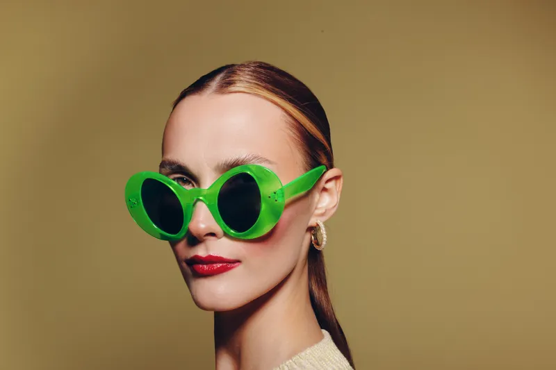 Stylish woman with slicked hair wearing large green sunglasses and red lipstick poses against beige background. Concept of fashion, accessories, and modern beauty portrait.