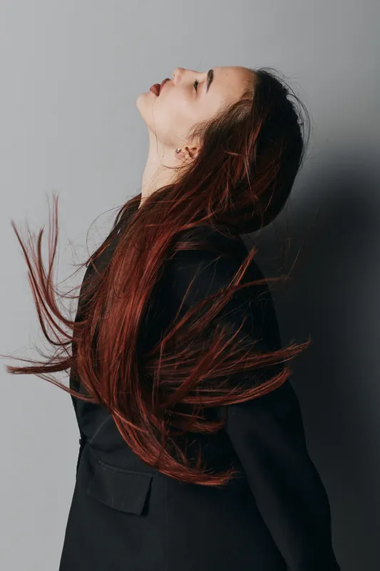 Woman with long red hair and closed eyes wearing black jacket standing against gray background. Side profile with flowing hair and serene expression in studio portrait.