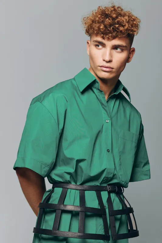 Young man with curly hair wearing green shirt and black cage belt posing against gray background in studio for fashion portrait and style concept.