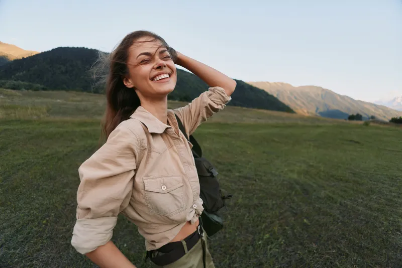 Cheerful woman outdoors, smiling with relaxed pose in a sunlit meadow, mountains in the background, natural colors, carefree lifestyle moment