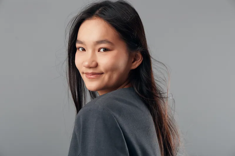 Young woman with long black hair smiling and turning head over shoulder on gray background with casual outfit and natural makeup for portrait or lifestyle concept.