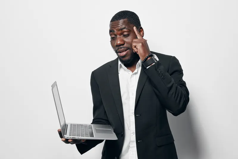 African American man with dark skin holding laptop and pointing to head in thoughtful gesture, wearing black suit and white shirt against white background.
