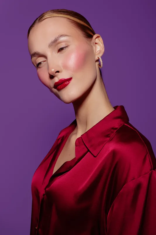 Portrait of a confident woman with glossy red lips and shiny skin wearing a red satin blouse against a purple background. Concept of beauty, fashion, elegance, and makeup.
