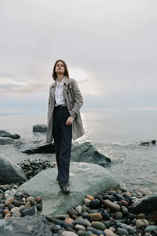 A confident woman in a patterned coat stands on rocky shore near calm sea under cloudy sky. Concept for fashion, confidence, nature, and outdoor lifestyle by the water.