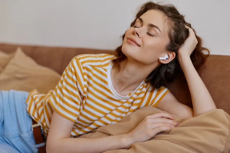 Young woman relaxes on a sofa wearing a yellow striped t shirt and blue jeans, listening to music with wireless earbuds, eyes closed, feeling cozy, content and at ease.