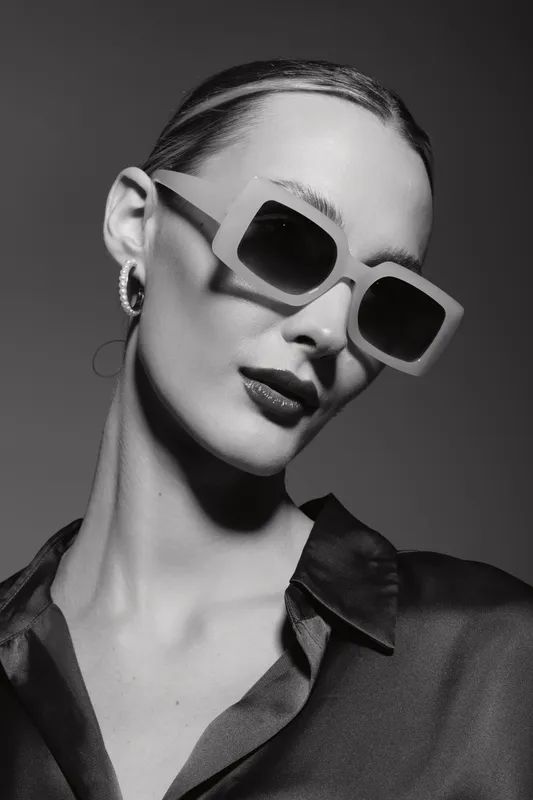 Elegant white woman with fair skin wearing large square sunglasses and hoop earrings poses in studio with dark shirt. Concept of fashion, style, and beauty portrait.