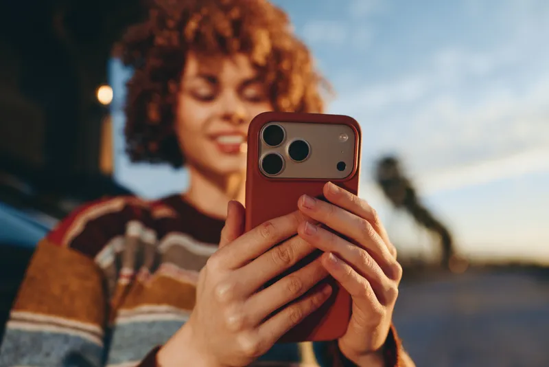 Woman lifestyle portrait outdoors at sunset holding smartphone for selfie, curly hair and relaxed smile, hands covering face slightly, golden evening light, casual street background with bokeh
