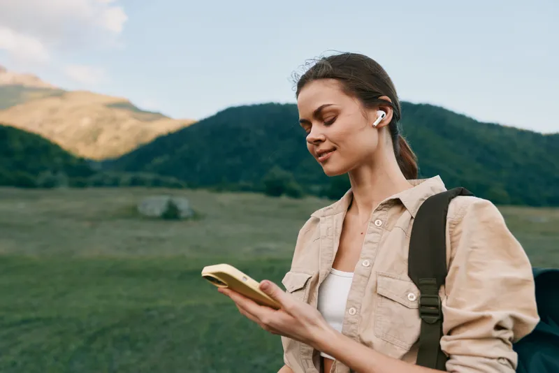 Young woman with a backpack listens to music on wireless earbuds while checking her smartphone outdoors in a mountainous area during daytime.