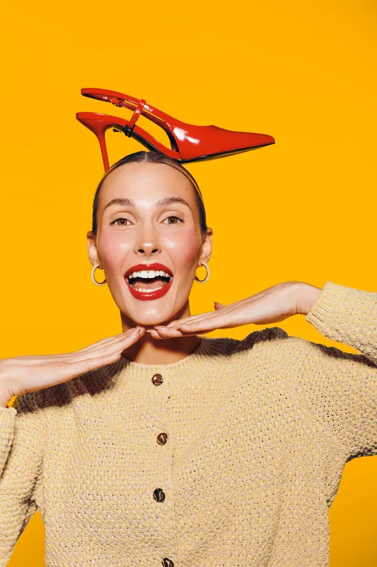 Happy woman with red high heel shoe balanced on head smiles brightly on yellow background. Concept for fashion, fun, creativity, and joyful lifestyle with vibrant colors and playful mood.