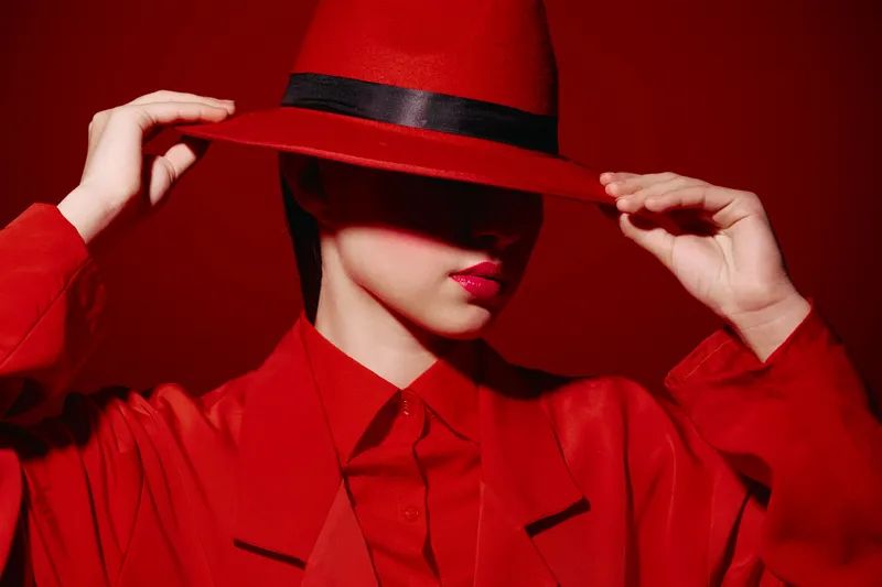 Fashionable woman in red outfit and hat partially covering face poses against red background. Artistic beauty photography emphasizing sophistication and style.