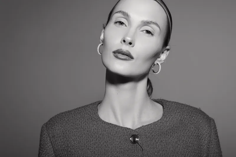 Portrait of a serious Caucasian woman with pale skin and slicked-back hair wearing elegant clothes and hoop earrings in black and white studio.