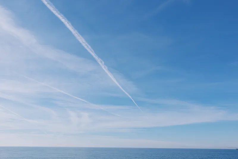 Bright blue sky over calm ocean with a distant horizon and a white contrail slicing through wispy clouds, creating a serene seascape ideal for travel and nature themes.