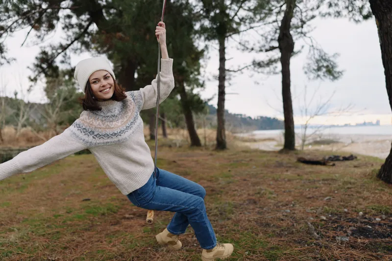 Girl on a swing in a cozy winter sweater and beanie, swinging by a lakeside park with pines and calm water, capturing playful movement and outdoor joy.