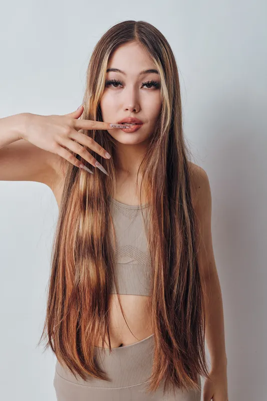 Woman with long hair and natural makeup posing with fingers near lips wearing beige crop top and swimsuit in studio with neutral background and soft lighting.