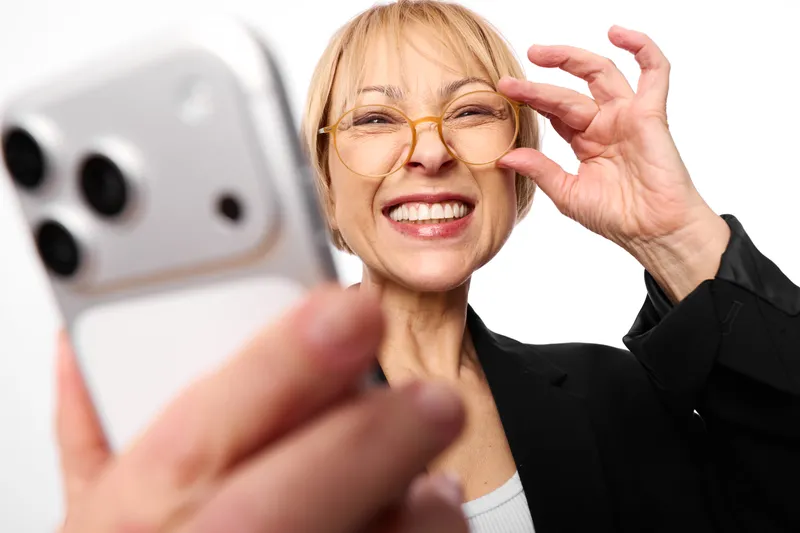 Woman with glasses smiles for selfie while holding smartphone, closeup portrait of senior, technology communication with joyful video cheerful vibe indoors today