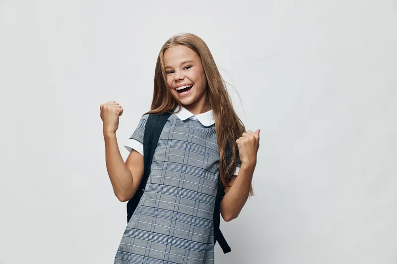 Girl with backpack smiling and raising fists in excitement standing on white background. Happy school student wearing uniform, preparation for education and learning.