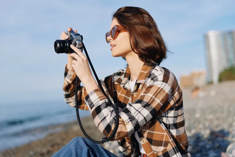 Woman photographer in plaid shirt with camera by the seaside, outdoor daylight, winding coast, clear sky, waves and rocks, capturing travel lifestyle moments with authentic creative energy