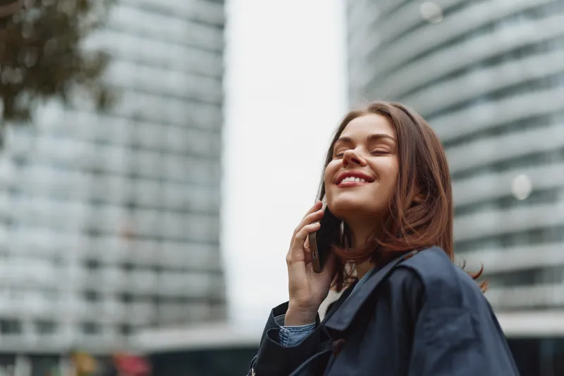 Smiling woman with dark hair on phone in an urban setting, wearing a dark coat, tall buildings behind her, a candid moment of confidence and connection.