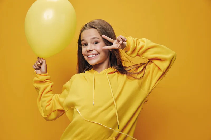 Girl smiling and holding yellow balloon with peace sign gesture for celebration and joy in studio. Happy child in yellow sweatshirt posing on yellow background.