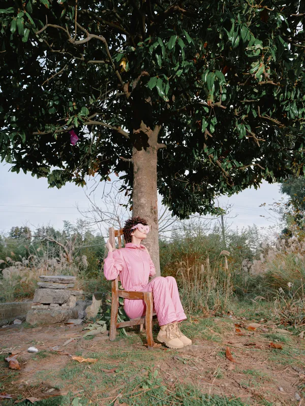 Woman in pink outfit sits on a wooden chair under a leafy tree in a rural outdoor setting, enjoying warm sunlight, rustic scenery and natural calm.