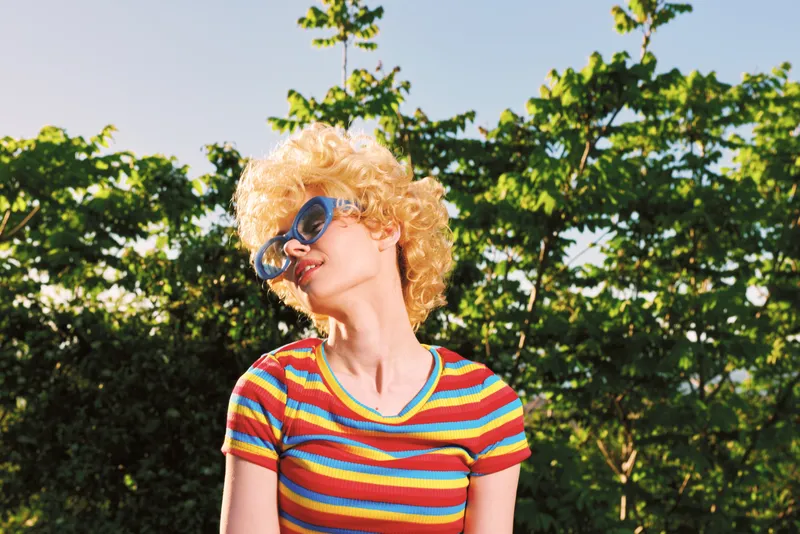 Cheerful woman outdoors, wearing a colorful striped shirt and oversized sunglasses, basking in sunlit greenery with a breezy vibe and retro charm and a timeless feeling of carefree summer style