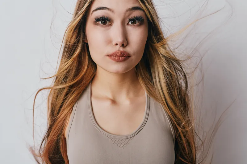Woman with long hair and neutral expression wearing beige tank top on white background. Portrait of young female with natural look and slight makeup.