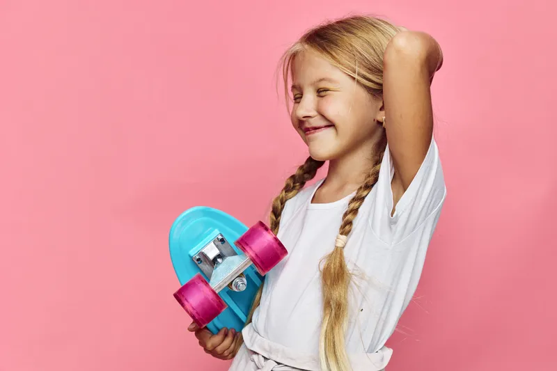 Smiling young girl with braided hair holding a blue skateboard against pink background. Casual outfit, natural light, happy expression, lifestyle concept.