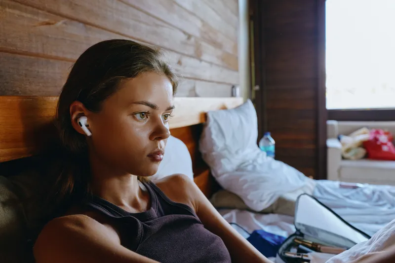 A woman relaxes on a bed in a cozy bedroom, wearing earbuds, listening to audio, capturing a casual lifestyle moment of comfort, rest, and quiet interior calm.