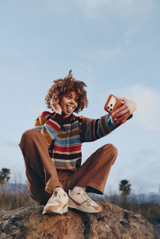 Smiling woman lifestyle selfie smartphone outdoors scene curly hair sitting on a rock at dusk she holds a smartphone for a selfie casual traveler mood natural light and clouds sky