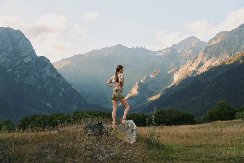 A young woman with long brown hair stands on a rock in a grassy field surrounded by mountains during golden hour, enjoying a moment of freedom and adventure.