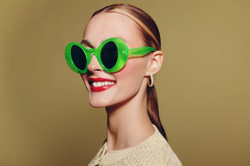 Smiling woman with slicked hair wears large green sunglasses and red lipstick. She poses against a beige background wearing a light sweater. Concept for fashion, style, and beauty.