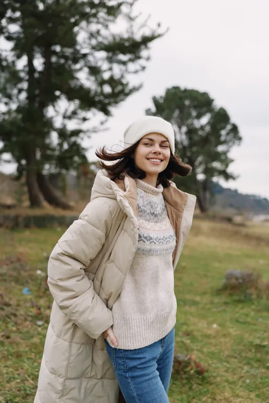 Casual outdoor portrait of a smiling woman in a warm coat and knit sweater, wearing a beanie, standing in a park with trees and soft natural light highlighting fashion and everyday comfort.