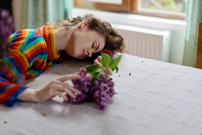 A woman rests her head on a table in a bright home, a bouquet of purple flowers nearby. A lifestyle scene conveying fatigue, calm, and moments of quiet recovery.